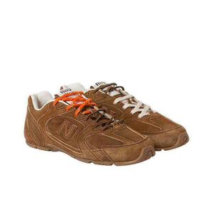 Pair of brown suede New Balance shoes shown at a three-quarter angle, featuring dual-lacing with contrasting orange and off-white laces, Miu Miu branded tongue tabs, and low-profile brown soles