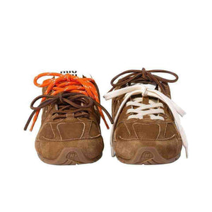 Straight-on toe-up view of brown suede New Balance shoes, displaying a multi-colored double lacing system with orange, brown, and off-white laces, co-branded tongue labels, and embossed "N" toe logos.