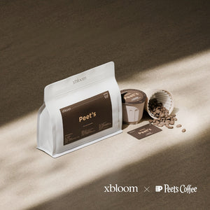 A white bag of Peet's Coffee with a dark brown label next to individual XBloom xpods coffee capsules and loose coffee beans, featuring the 'xbloom x Peet's Coffee' collaboration logo.
