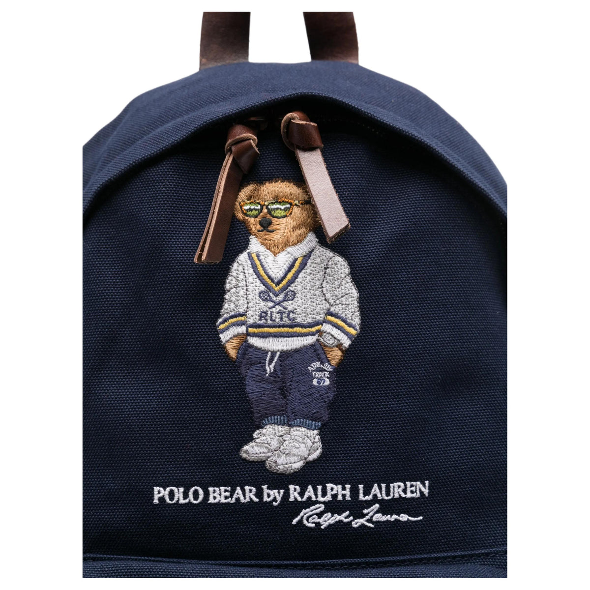 Close-up of the iconic embroidered Polo Bear wearing a sweater and glasses on an original navy Ralph Lauren backpack with brown leather zipper pulls.

لقطة قريبة لتطريز دب بولو الشهير الذي يرتدي سترة ونظارات على حقيبة ظهر رالف لورين أصلية باللون الكحلي مع أشرطة سحاب جلدية بنية.